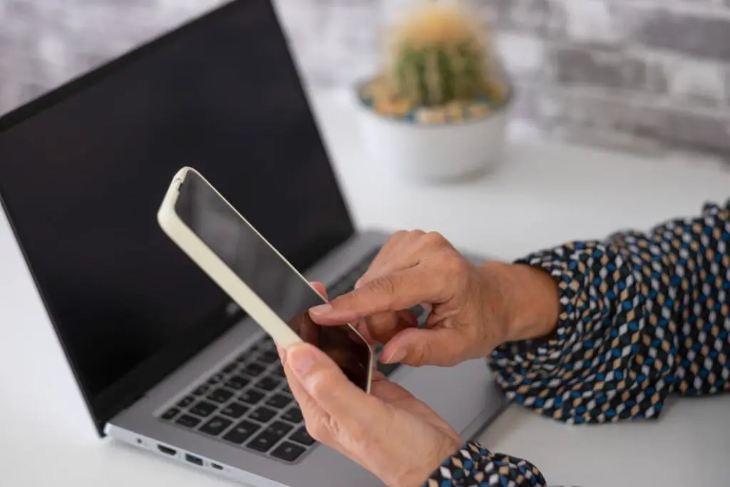 An image of a woman hands using mobile phone while working with laptop, mature female browsing site on smartphone.