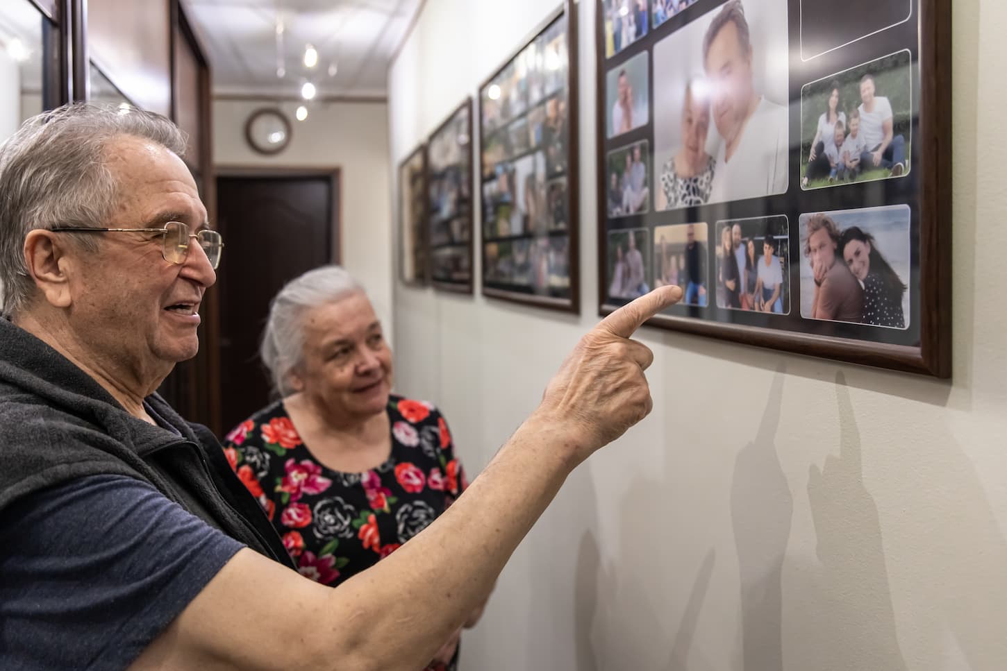 An image of grandparents looking at family photographs on the wall in their home.